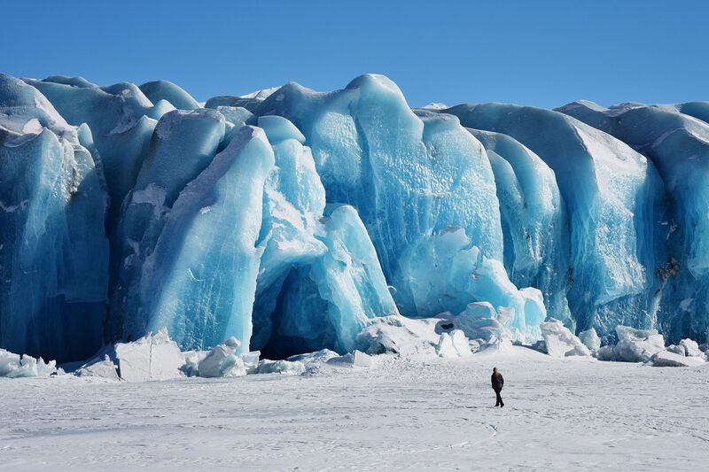 A person walking on the frozen lake below the blue ice face of Knik Glacier